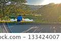 A person sits by the roadside, taking in the beautiful vista of a lake and mountains bathed in golden sunlight 133424007
