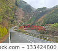 A vibrant red bridge spans a river, with a road leading through a verdant valley surrounded by mountains 133424600