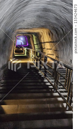 A dimly lit tunnel with stone stairs and metal railings leads upward toward a bright exit.  133424673