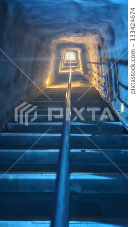 A dimly lit tunnel with stone stairs and metal railings leads upward toward a bright exit.  133424674
