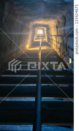 A dimly lit tunnel with stone stairs and metal railings leads upward toward a bright exit. A dimly lit tunnel with stone stairs and metal railings leads upward toward a bright exit. 133424675