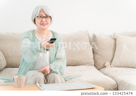 A senior woman with gray hair sits on the living room sofa and operates the TV remote control. A senior woman with gray hair sits on the living room sofa and operates the TV remote control. 133425474