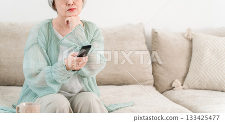 A senior woman with gray hair sits on the living room sofa and operates the TV remote control. A senior woman with gray hair sits on the living room sofa and operates the TV remote control. 133425477