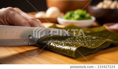 Professional chef hands slicing fresh green kelp seaweed with sharp knife on wooden cutting board in modern kitchen with water droplets 133427224