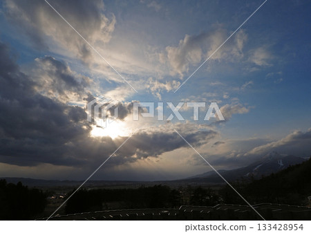 The sun shines through the gaps in the clouds at the base of Mt. Bandai. The radiating light shines onto the ground, creating an impressive and beautiful contrast with the blue sky. 133428954