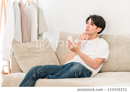 一位年輕的亞洲男子坐在客廳沙發上，一邊看著他的智慧型手機一邊思考。 133430352