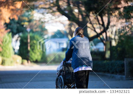 A woman caring for an elderly person in a wheelchair outdoors in autumn among fallen leaves. Wide-angle, full-body, faceless back view. Visiting care, walking, etc. A woman caring for an elderly person in a wheelchair outdoors in autumn among fallen leaves. Wide-angle, full-body, faceless back view. Visiting care, walking, etc. 133432321
