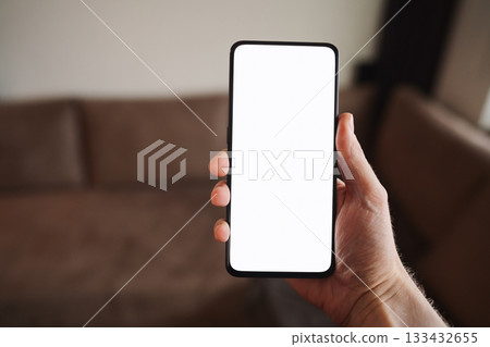 Man hold smartphone with white screen in his hand standing indoors in home environment Man hold smartphone with white screen in his hand standing indoors in home environment 133432655