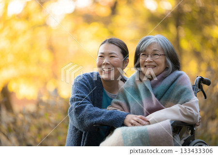 一位老婦人坐在輪椅上，漫步在美麗的秋葉間，身邊有一位護理人員和一位看起來像是她女兒的人。 133433306