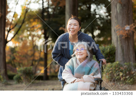 一位老婦人坐在輪椅上,漫步在美麗的秋葉間,身邊有一位護理人員和一位看起來像是她女兒的人。 一位老婦人坐在輪椅上,漫步在美麗的秋葉間,身邊有一位護理人員和一位看起來像是她女兒的人。 133433308