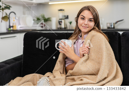 Young woman with tea cup warms hands wraps in blanket squints and enjoys calm serene evening tonight 133433842