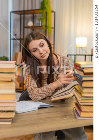 Young woman surrounded by books writes notes from textbook feels exhausted yet continues heavy study Young woman surrounded by books writes notes from textbook feels exhausted yet continues heavy study 133433854
