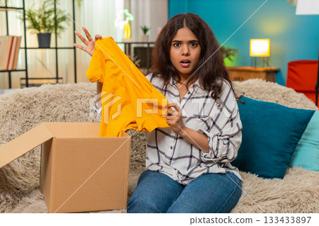 Indian woman unboxes package find wrong t-shirt frowns disappointed shakes head online shopping flaw 133433897