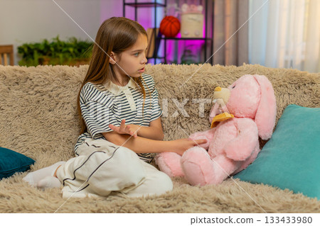 Child girl kid talking to plush bunny toy while sitting on home sofa in playful and imaginative mood Child girl kid talking to plush bunny toy while sitting on home sofa in playful and imaginative mood 133433980