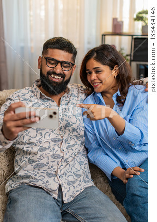 Indian family couple man woman laughing together at funny videos on smartphone sitting on home sofa Indian family couple man woman laughing together at funny videos on smartphone sitting on home sofa 133434146