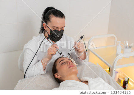 Beautician doctor makes woman micro currents face therapy using electrical impulses in spa salon 133434155