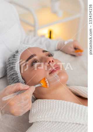 Enzyme therapy, beautician applying moisturizing medical cream on young woman face with brushes 133434156