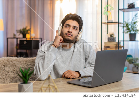 Young Indian man wearing wireless headphones working on laptop while listening to audiobook at home 133434184