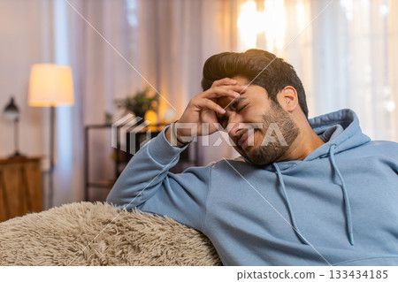 Indian young man on sofa rubbing temples to cure headache suffering from stress and migraine at home 133434185