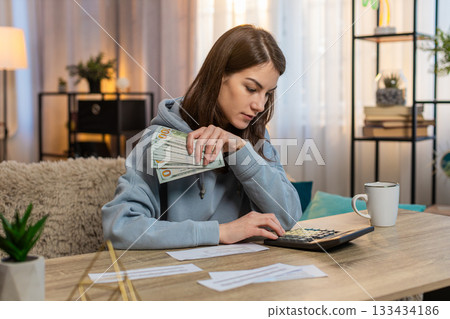 Young woman counting money while sitting on sofa with receipts and calculator on tablet at home Young woman counting money while sitting on sofa with receipts and calculator on tablet at home 133434186