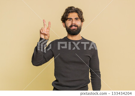 Young Indian man showing victory sign, doing peace gesture standing against beige studio background Young Indian man showing victory sign, doing peace gesture standing against beige studio background 133434204