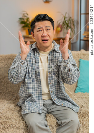 Upset Chinese man making face disappointed in result, bad news watching TV on sofa couch at home 133434214