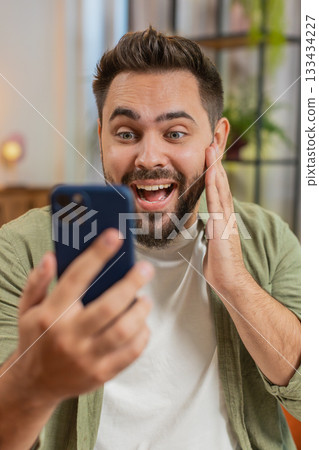 Surprised Caucasian brunette man using smartphone winning lottery sitting on sofa couch at home 133434227