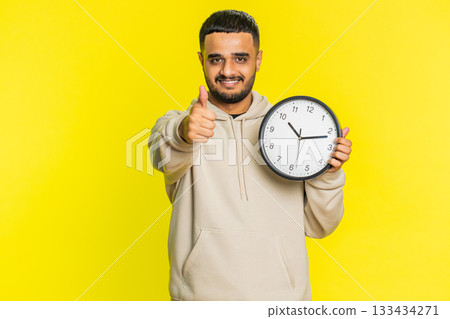Indian young man showing time hour on wall office clock, ok, thumb up, approve, hurry up, deadline Indian young man showing time hour on wall office clock, ok, thumb up, approve, hurry up, deadline 133434271