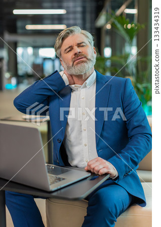 Tired middle-aged businessman suffers from spine neck muscle pain working on laptop in office lobby 133434319