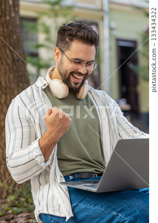 Overjoyed happy young man working on laptop netbook screaming in delight in urban city street Overjoyed happy young man working on laptop netbook screaming in delight in urban city street 133434322