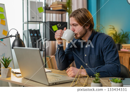 Businessman working on laptop drinking cup of warm hot coffee sits at table workplace home office 133434331