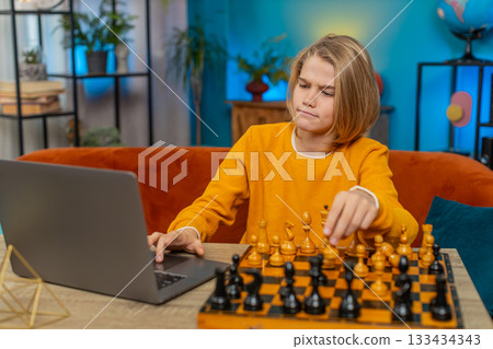 Teenager boy playing online chess on laptop with real chessboard thoughtfully moving pieces at table Teenager boy playing online chess on laptop with real chessboard thoughtfully moving pieces at table 133434343
