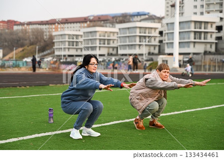 Two Russian women, aged 50 and 60, in tracksuits,doing squats at the stadium 133434461