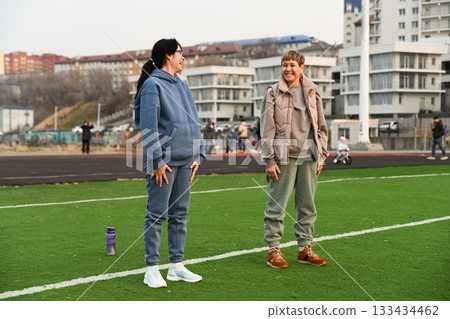 Two Russian women, aged 50 and 60, in tracksuits,doing squats at the stadium and laughing Two Russian women, aged 50 and 60, in tracksuits,doing squats at the stadium and laughing 133434462