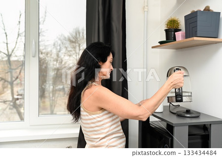 A 50 year old Russian woman with long dark hair prepares hot water for morning coffee, still sleepy, standing by the window in a calm cozy kitchen atmosphere. 133434484