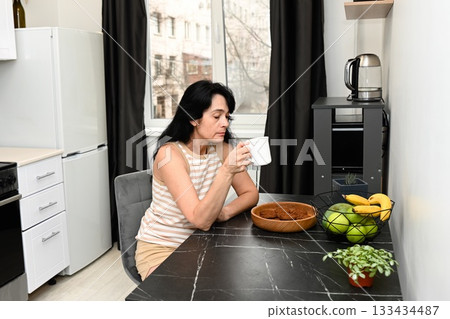 Mature 50 years old Russian woman sits with a cup of coffee on a calm sleepy morning, looking thoughtful and relaxed in a soft peaceful mood. Mature 50 years old Russian woman sits with a cup of coffee on a calm sleepy morning, looking thoughtful and relaxed in a soft peaceful mood. 133434487