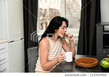 Mature 50 years old Russian woman enjoys a calm sleepy morning holding coffee and a cookie, taking a bite while sitting quietly with a soft peaceful mood. Mature 50 years old Russian woman enjoys a calm sleepy morning holding coffee and a cookie, taking a bite while sitting quietly with a soft peaceful mood. 133434489