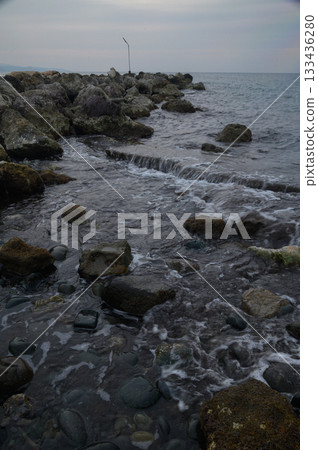 Waves gently lap against rocky shore while distant tree stands alone edge of the water at twilight Waves gently lap against rocky shore while distant tree stands alone edge of the water at twilight 133436280