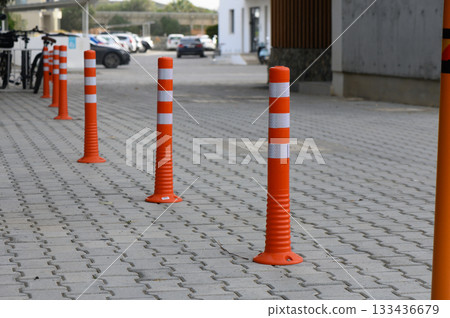 Bright orange safety bollards line a pedestrian walkway in a modern urban setting during daylight hours Bright orange safety bollards line a pedestrian walkway in a modern urban setting during daylight hours 133436679