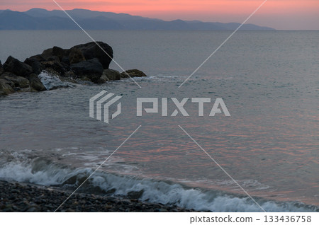 Serene sunset over tranquil waters with gentle waves lapping at rocky shorelines in a coastal landscape 133436758