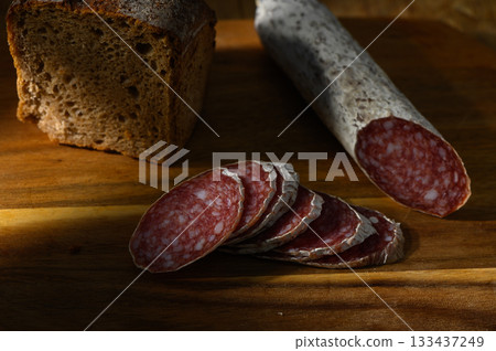 Slices of cured salami beside hearty bread on a wooden cutting board 133437249