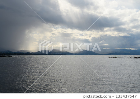 Dramatic clouds cast shadows over tranquil waters near distant mountains during a cloudy afternoon Dramatic clouds cast shadows over tranquil waters near distant mountains during a cloudy afternoon 133437547