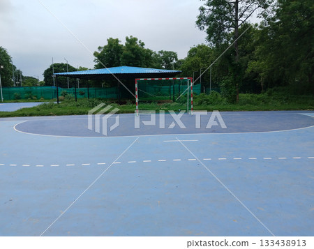 Outdoor futsal goalpost with striped metal frame and loose green net on blue sports court, standalone mini football goal in open ground surrounded by grass and trees, dedicated small-sided play area Outdoor futsal goalpost with striped metal frame and loose green net on blue sports court, standalone mini football goal in open ground surrounded by grass and trees, dedicated small-sided play area 133438913