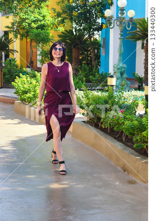 A woman in a beautiful cherry-colored dress walks dancing through a neighborhood with expensive houses 133440300