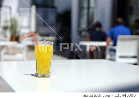 Glass of orange juice with plastic straw on white table in restaurant outdoor lounge zone in sunny weather Glass of orange juice with plastic straw on white table in restaurant outdoor lounge zone in sunny weather 133440390
