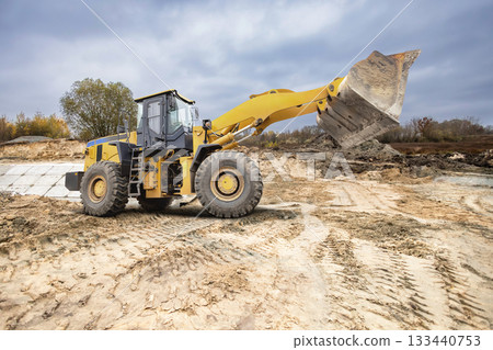 A large yellow loader works to move earth at a construction site surrounded by trees during cloudy autumn weather. Dust rises with each scoop 133440753