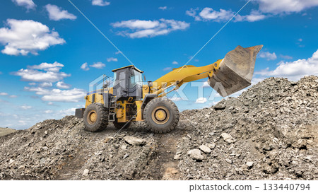 A large yellow loader is seen moving gravel at a construction site, surrounded by heaps of rocks and under a beautiful blue sky A large yellow loader is seen moving gravel at a construction site, surrounded by heaps of rocks and under a beautiful blue sky 133440794