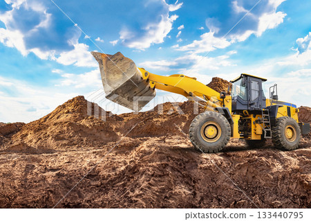 A yellow A wheeled bulldozer or loader lifts a large scoop of dirt at a construction site. The bright sky and piles of earth create a busy atmosphere in the daytime 133440795