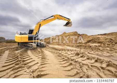 A yellow excavator digs into the sandy ground at a construction site with piles of earth under a cloudy sky during the afternoon 133440797