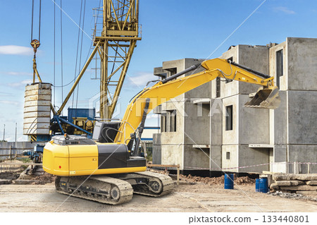 Excavator lifts material at a construction site with cranes and concrete structures in the background, showcasing urban development during the day 133440801